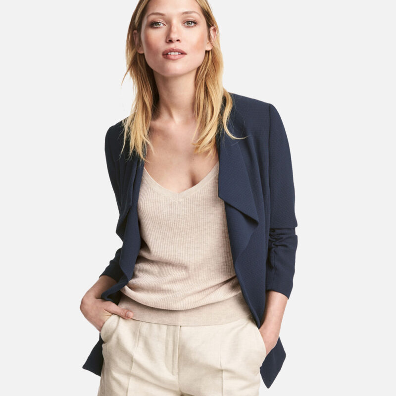 Blonde woman in beige top and navy jacket with hands in pockets.